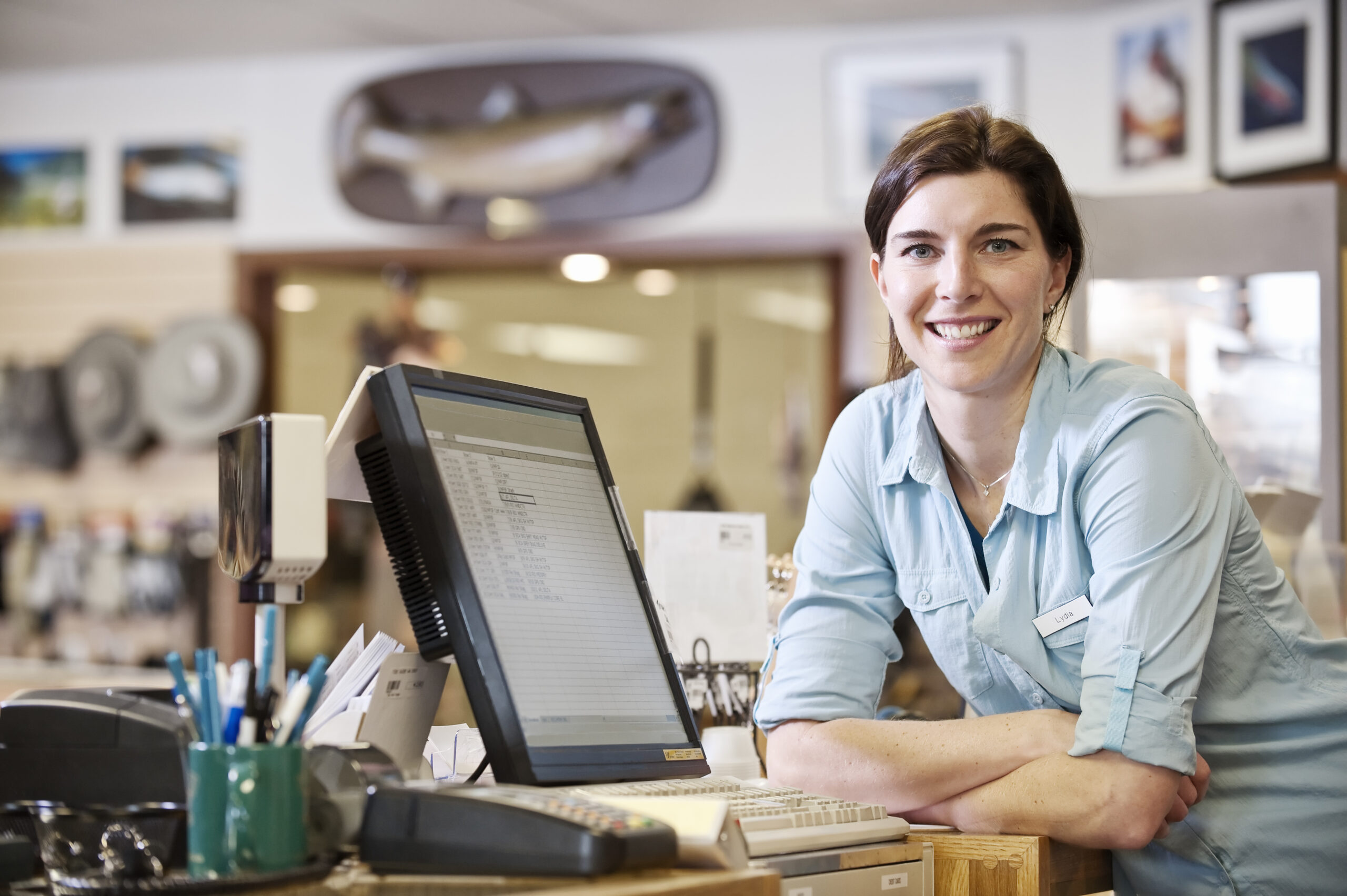 Portrait of a female caucasian owner next to the computer at the front desk of a fly-fishing shop.